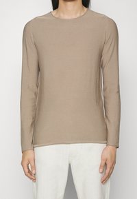 T-shirt en coton marron clair à manches longues avec un col rond et une texture lisse, associé à un pantalon écru.