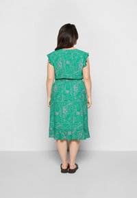 Vestido verde com mangas curtas e volumosas, padrão floral em rosa, cintura ajustada e comprimento até o joelho. Usado com sandálias pretas com costas abertas.
