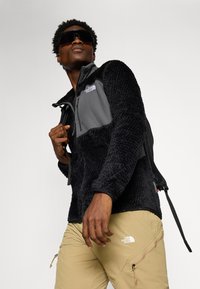 Fleecejack in zwart met grijze panelen, ritszak en textuurafwerking, gecombineerd met khaki broek met ritsen en logo. Zonnebril op.