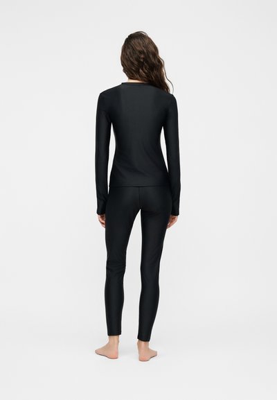 Γυναίκα που στέκεται ξυπόλητη με την πλάτη γυρισμένη, φορώντας εφαρμοστή μαύρη μακρυμάνικη μπλούζα και ταιριαστό μαύρο κολάν σε απλό φόντο.