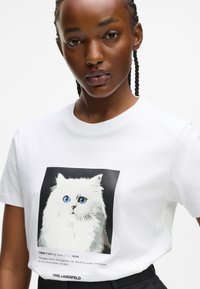 Valge puuvillane T-särk, millel on graafiline print karvases valges kassist siniste silmadega, musta tausta ees, ja tekst all.