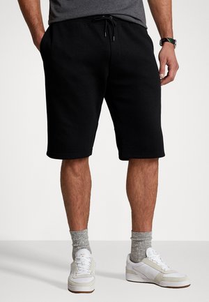 Mann trägt schwarze Shorts mit Kordelzug, graue Knöchelsocken, weiße Sneaker und ein graues Shirt, steht mit entspannten Händen an den Seiten.