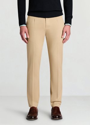 Pantaloni beige su misura con una trama liscia e pieghe anteriori, abbinati a scarpe eleganti marroni. La maglia è nera a costine con polsini.