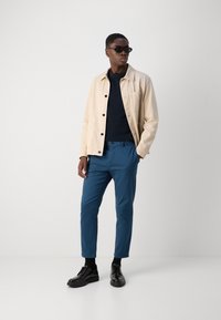 Beige dzseki egy sötétkék pulóver felett, kék nadrággal és fekete cipővel párosítva. A modell napszemüveget visel és laza testtartásban áll.