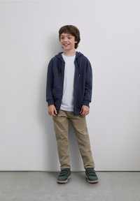 Hanorac bleumarin cu fermoar, tricou alb, pantaloni bej și pantofi verzi. Material neted, design casual, cu buzunare frontale pe hanorac.