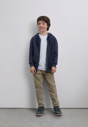 Sudadera con cremallera azul marino, camiseta blanca, pantalones color caqui y zapatos verdes. Tela suave, diseño casual, con bolsillos delanteros en la sudadera.