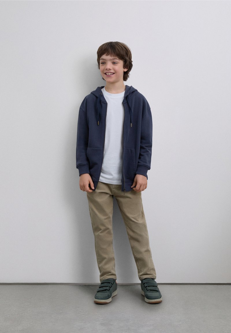 Hanorac bleumarin cu fermoar, tricou alb, pantaloni bej și pantofi verzi. Material neted, design casual, cu buzunare frontale pe hanorac.