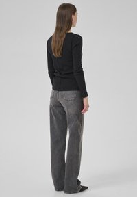 Woman with long brown hair wearing black long-sleeve top, loose gray jeans, and black pointed shoes, standing against a plain light gray background.