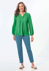 Blusa verde a maniche lunghe con scollo a V e dettagli plissettati, abbinata a pantaloni blu a fantasia con forme geometriche. Sandali completano l'outfit.