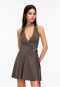 Brown halter dress with a deep V-neck, wrap design, and a silver loop accent. Flared skirt with tie detail on the side. Soft, smooth fabric.