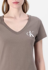 Rudas medvilninis marškinėliai su V formos iškirpte ir trumpomis rankovėmis, kuriuose kairėje krūtinės pusėje yra balta Calvin Klein Jeans logo.