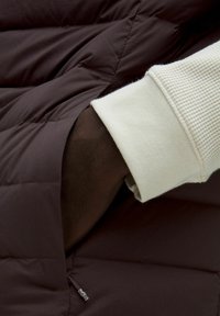 Giacca a vento marrone con una tasca zip, che mostra un polsino in maglia bianca testurizzata sotto. Il tessuto appare liscio e isolato.