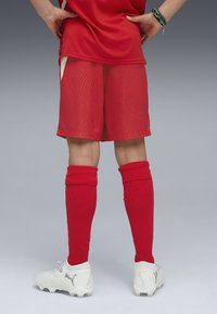 Pantalones cortos de fútbol rojos con textura acanalada vertical, combinados con calcetas rojas hasta la rodilla y tacos de fútbol blancos con parte superior texturizada y logotipo.