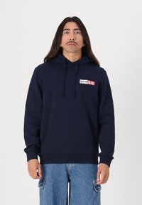 Mørkeblå hettegenser med en frontlomme, med en rød og hvit logo-patch. Laget av bomull, avslappet passform, ribbestrikkede mansjetter og hem.