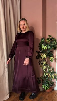 Γυναίκα με μακρύ σκούρο μοβ σατέν φόρεμα στέκεται κοντά σε μπεζ κουρτίνες και ένα πολύχρωμο πράσινο φυτό σε ξύλινο πάτωμα σε ένα ροζ δωμάτιο.