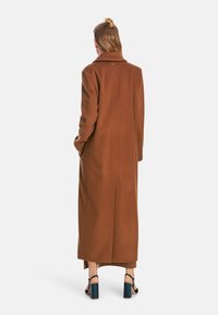 Long brown wool coat featuring a notched collar, side pockets, and a loose fit, paired with teal-colored heeled sandals.