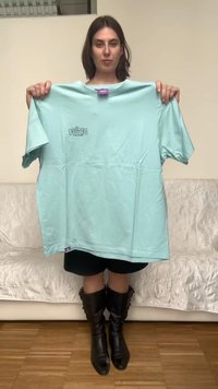 Kobieta stoi w pomieszczeniu, trzyma jasnoniebieski, oversize'owy t-shirt z małym napisem na piersi, nosi czarne szorty i czarne, sięgające kolan kozaki.