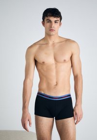 Boxeri negri cu un tiv elastic roșu și albastru, purtând numele mărcii "HUGO". Materialul pare neted și elastic.