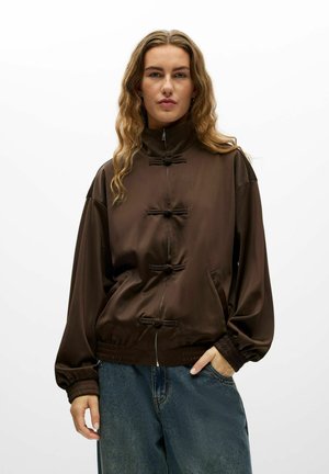Personne aux longs cheveux ondulés portant une veste marron zippée avec des détails de boutons en boucle et un jean bleu, debout devant un fond blanc uni.