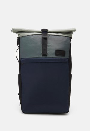 Mochila de rollo en tela estratificada; presenta secciones en gris, verde azulado y azul marino con una base negra. Incluye un bolsillo frontal con cremallera y una correa ajustable.