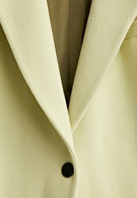 Blazer en mélange de laine jaune clair avec un col cranté, une fermeture à un bouton noir et un tissu doux texturé. L'intérieur est d'un beige discret.