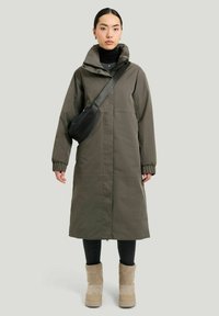 Lang olivengrøn parka med høj krave, elastiske manchetter og lige snit. Kombineret med sorte leggings og beige ankelstøvler.