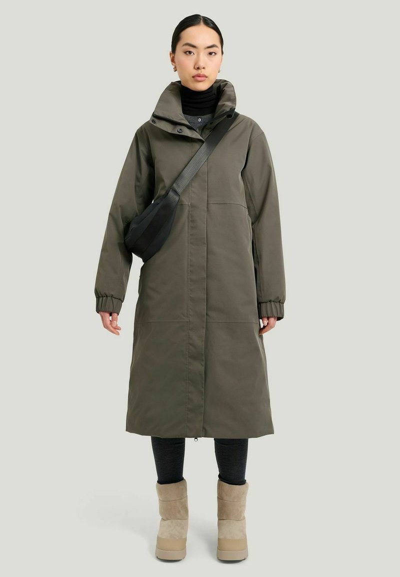 Lång olivgrön parka med hög krage, elastiska manschetter och rak skärning. Kombinerad med svarta leggings och beige ankelstövlar.