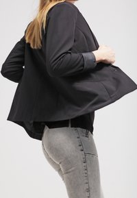 Osoba upravující černý blejzr a přitom nosící světle šedé upnuté džíny, viděná z boku na pozadí s jednoduchým světlým tónem.