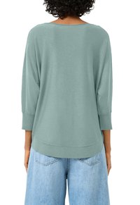 Kobieta z kręconymi włosami, ubrana w luźną, jasnoniebiesko-zieloną bluzkę z długim rękawem oraz jasnoniebieskie dżinsy, pokazana od tyłu.