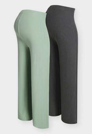 Pikkade mustade juustega naine, kes kannab musta varrukateta tärklisega ülaosa ja kõrge vöökohaga halli laiade säärtega pükse, hoides musta käekotti, seisab.