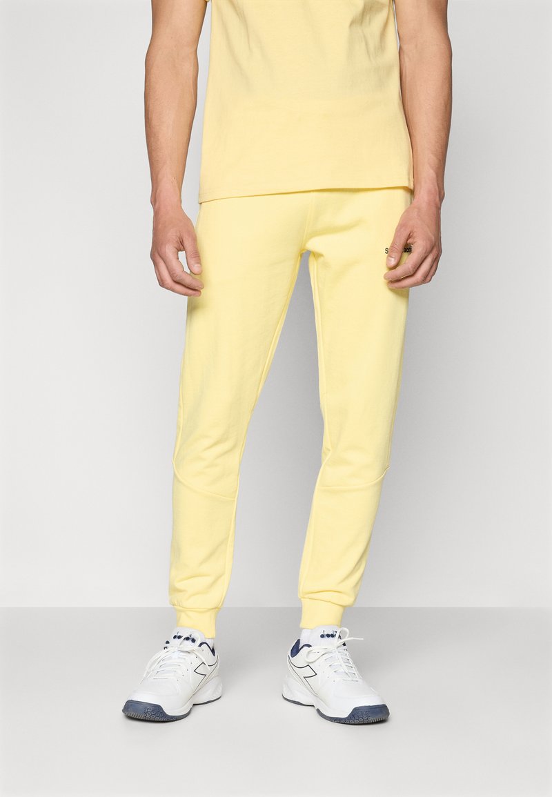 T-shirt en coton jaune associé à un jogging jaune assorti, avec une coupe ajustée et des poignets côtelés. Des baskets blanches complètent le look.