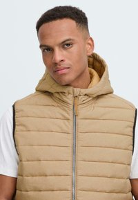Chaleco acolchado beige con capucha, que cuenta con rayas horizontales, un cierre frontal y acentos oscuros en contraste en los hombros.
