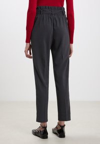 Pantaloni su misura grigio scuro con una vita alta e arricciata. Texture liscia, vestibilità slim e lunghezza alla caviglia, abbinati a scarpe basse nere.