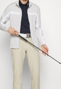 Vaaleanharmaa vetoketjullinen takki, jossa valkoiset yksityiskohdat, päällä tummansininen pikeepaita. Beige housut ja valkoinen vyö, golfmaila kädessä. Sileä kangaspinta.