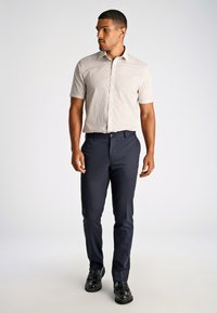 Camicetta a maniche corte con motivo nei toni del beige abbinata a pantaloni blu scuro. Il modello indossa scarpe nere, mostrando una silhouette dalla vestibilità rilassata.