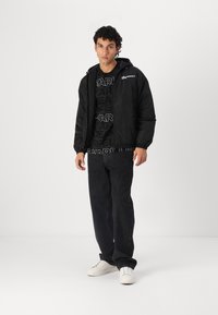 Black padded jacket with a hood, elastic cuffs, and branding. Worn over a black patterned shirt and paired with dark jeans and white sneakers.