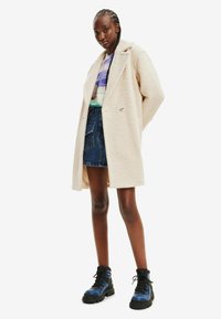 Noor naine kannab beeži tekstuuriga mantlit, mustriga särki, teksaseelikut ja siniseid lahingusaapaid, seistes valge tausta ees.