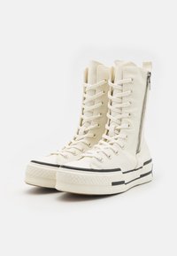 White high-top sneakers made of canvas with a side zipper, round toe, and black rubber sole featuring two black stripes for contrast.