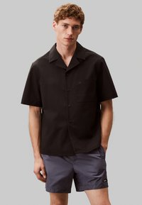 Camisa negra de manga corta con botones y un acabado texturizado, presenta un bolsillo en el pecho y un logo sutil. Combinada con pantalones cortos gris oscuro.