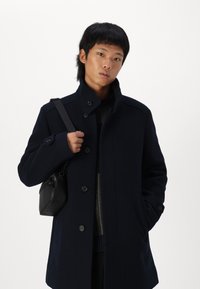 Navy wool blend coat featuring a high collar, button closure, side pockets, and a structured design. Paired with a small black bag.