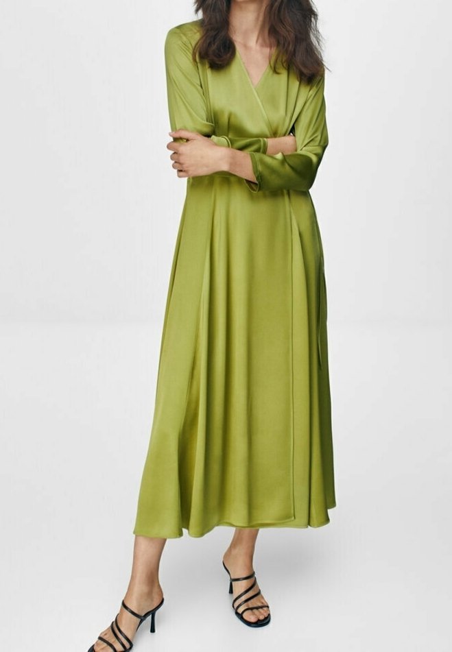 Femme portant une longue robe portefeuille soyeuse vert olive à manches longues et des sandales à talons hauts noires à brides, debout les bras croisés.