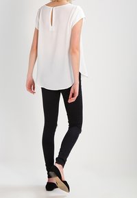 Blusa bianca a maniche corte con dettaglio cut-out sulla schiena, abbinata a jeans neri skinny e ballerine nere, con una superficie di tessuto liscia.