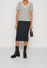 T-shirt a righe bianche e nere con scollo a V abbinato a una gonna midi blu navy a costine e stivali neri alla caviglia, mentre tiene una piccola borsa bordeaux.