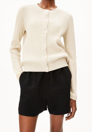 Cardigan côtelé beige avec un col rond, comportant six boutons à l'avant et des manches longues, associé à un short noir. Texture douce, design décontracté.