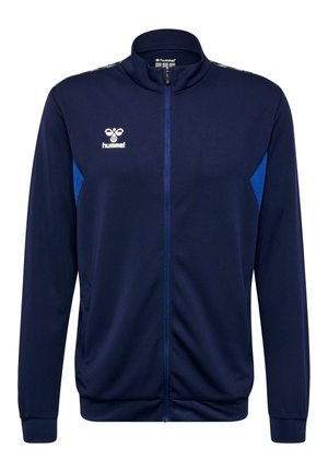 Marineblauwe sportjacket van gestructureerde stof, met een volledige ritssluiting, hoge kraag en blauwe accenten aan de zijkanten. Hummel-logo op de borst.