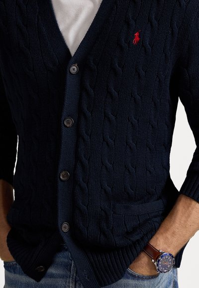 Καστόρινο ναυτικό πλεχτό ζακέτα με V-λαιμόκοψη, πέντε κουμπιά και δύο μπροστινές τσέπες, συνδυασμένο με ρολόι σε καφέ δερμάτινο λουράκι.