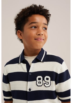 Garçon aux cheveux bouclés portant un polo rayé bleu marine et blanc avec le numéro 89, regardant légèrement sur le côté et souriant doucement.