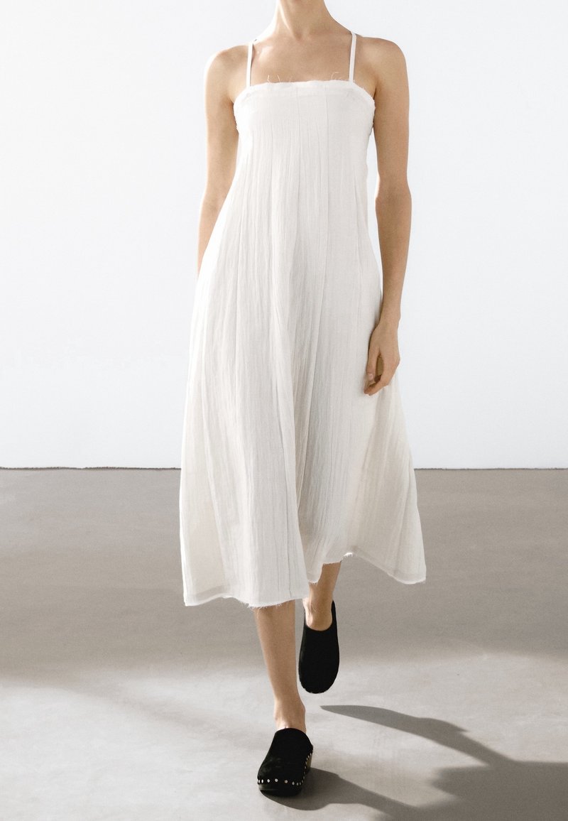 Robe blanche sans manches, longue jusqu'aux chevilles, en tissu texturé et fluide. Accompagnée de chaussures noires à semelle plate avec des clous argentés.