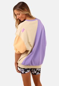 Pull en couleurs pastel—jaune, lavande et pêche—avec des manches en ballon et un patch logo texturé sur l'épaule.