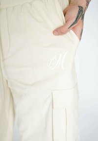 Beige cargo bukser har en afslappet pasform, elastisk talje, sidelommer og et hvidt broderet "M" logo på låret.